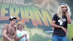 Mann mit langer blonder Perücke singt, zwei Männer im Hintergrund singen mit