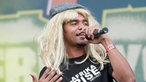 Mann mit langer blonder Perücke und Mütze singt in ein Mikro