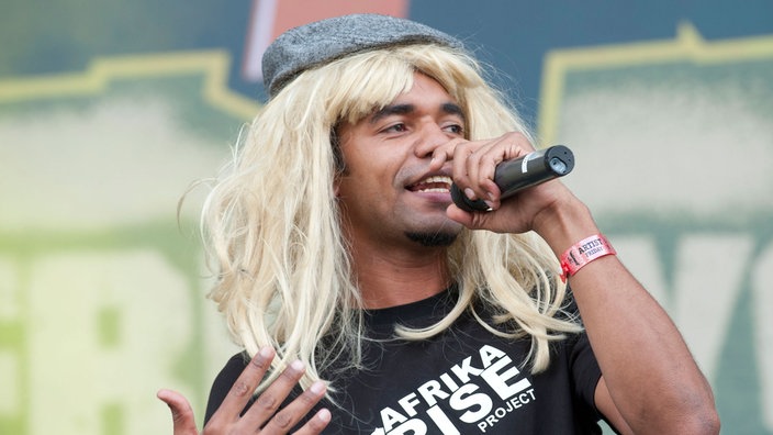 Mann mit langer blonder Perücke und Mütze singt in ein Mikro