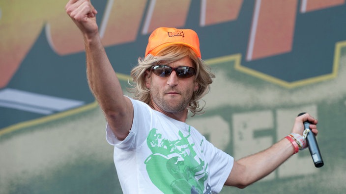 Mann mit blonder Perücke, orangem Hut und Sonnenbrille tanzt mit Mikrofon in der Hand