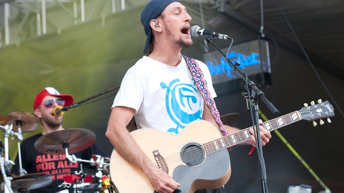 Mann mit Akustikgitarre und umgedrehter Kappe singt in ein Mikro.