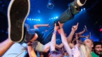 Stimmungsvolle Nahaufnahme eines Stage-Divers, der vom Publikum unter ihm getragen wird. Besonders nah sieht man die Sohle seines einen Turnschuhs, das andere Bein grätscht er in eine andere Richtung weg.