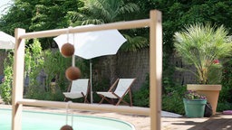 Im Hintegrund ein Garten mit Pool und Liegestühlen, im Vordergrund steht ein Outdoor-Spiel aus einem Holzgestell.
