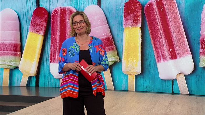 Yvonne Willicks steht im Serviczeit-Studio. Im Hintergrund sieht man ein Bild mit mehreren Eis am Stiel.