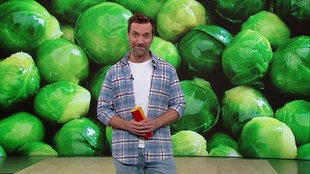  Daniel Aßman steht im Servicezeit-Studio. Im Hintergrund sieht man eine Nahaufnahme eines Dutzen Rosenkohls.