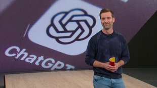  Daniel Aßmann steht im Servicezeit-Studio. Im Hintergrund sieht man das App-Logo der der KI "ChatGPT".