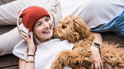 Eine lachende Frau mit roter Mütze und Kopfhörern kuschelt mit einem Hund 