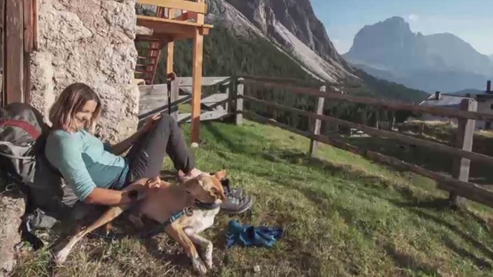 Eine Frau und ein Hund liegen auf einer Wiese mit Alpenpanorama im Hintergrund 