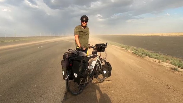 Ein Mann steht mit seinem Fahrrad auf einer Straße und trägt einen Fahrradhelm 