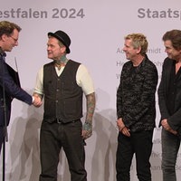 Fünf Männer auf einer Bühne, zwei davon  schütteln sich die Hand