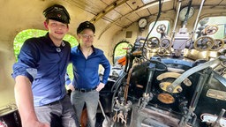 Zwei Männer in Uniform stehen im Führerhaus einer alten Dampflokomotive.