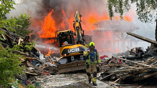 Ein Feuerwehrmann läuft vor einem Bagger, im Hintergrund stehen Trümmer in Flammen.