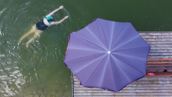 Eine Frau im Badeanzug schwimmt im Wasser. Ein großer Sonnenschirm steht daneben. 