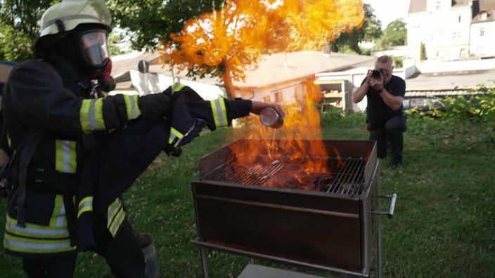 Ein Feuerwehrmann kippt Brandbeschleuniger auf die Glut in einem Grill und eine hohe Flamme schlägt ihm entgegen