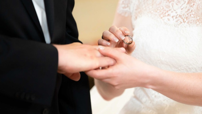 Eine Frau im weißen Kleid hält einen Ring in der Hand, in der anderen hält sie die Hand eines Mannes, der seine Finger ausstreckt, der Mann trägt Schwarz