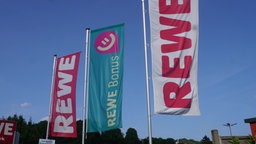 Das Bild zeigt REWE Fahnen im Wind. Darunter in der Mitte auch eine Fahne mit einer Werbung für die REWE Bonus-App.