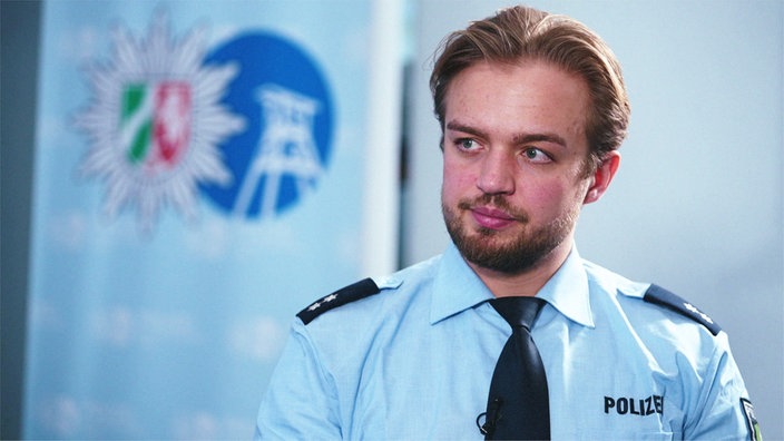 Hendrik Heyer, ein junger blonder Mann mit kurzen Haaren im Mittelscheitel und Vollbart, sitzt im blauen Hemd vor einer hellen Wand und schaut nach links aus dem Bild.