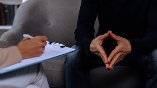 Zwei Personen sitzen sich gegenüber, die eine hält ein Klemmbrett in der Hand, der andere macht mit den Fingern eine Raute.