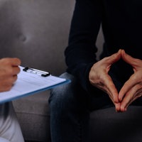 Zwei Personen sitzen sich gegenüber, die eine hält ein Klemmbrett in der Hand, der andere macht mit den Fingern eine Raute.