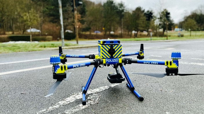 Auf der Straße steht eine Drohne. Sie ist blau und gelb mit der Aufschrift "Polizei".