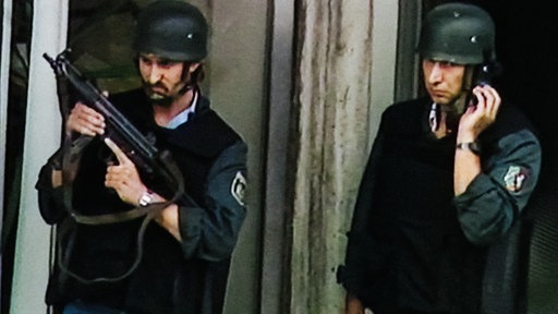 Zwei Polizisten in Schutzausrüstung stehen an der Sparkasse. Einer hält eine Schusswaffe in der Hand.
