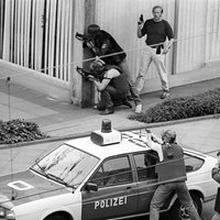 Polizisten zielen mit Waffen um die Ecke des Bankgebäudes, zwei weitere über ein Polizeiauto hinweg.