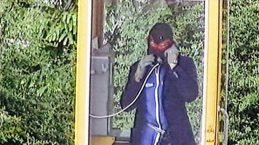 Einer der Täter telefoniert maskiert in einer Telefonzelle.