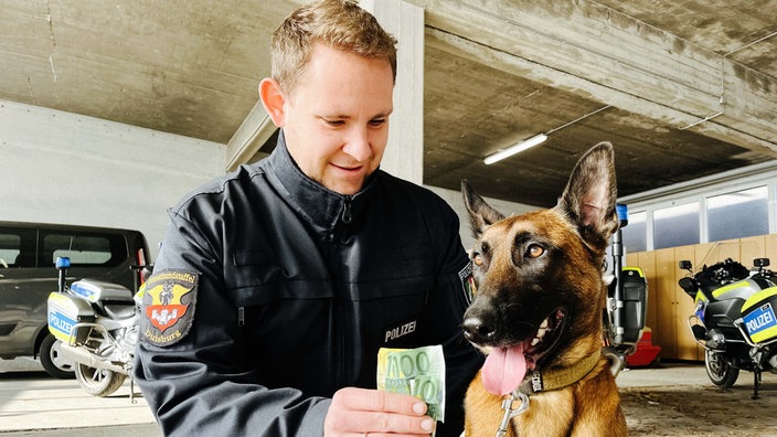 Gregor Köpp kniet in Unifrom neben dem Spürhund Cash. In der Hand hält er mehrere Geldscheine, die er neben den Kopf von Cash hält. Hinter ihnen sind zwei Polizeimotorräder. 