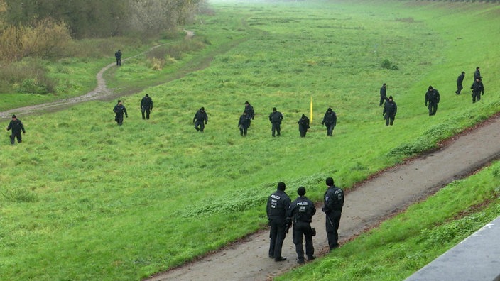 Auf einer grünen Wiese laufen zahlreiche schwarzgekleidete Polizeibeamte und suchen nach Spuren