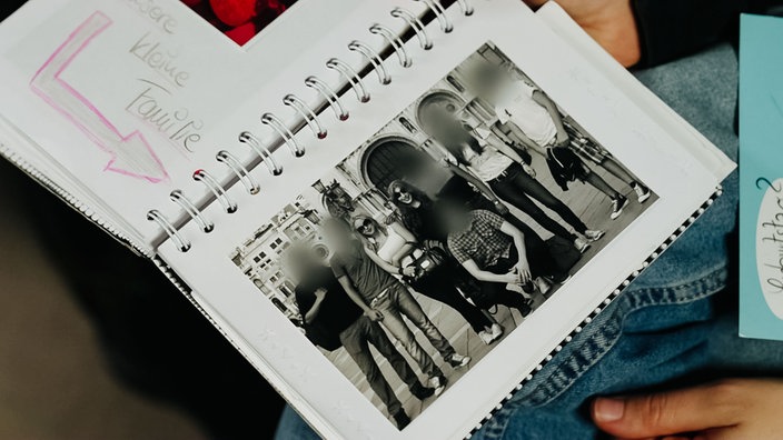 In einem Fotoalbum klebt ein Foto einer Freundesgruppe, in der Mitte stehen Derya Seyhun und Saskia, die restlichen Gesichter sind verpixelt.
