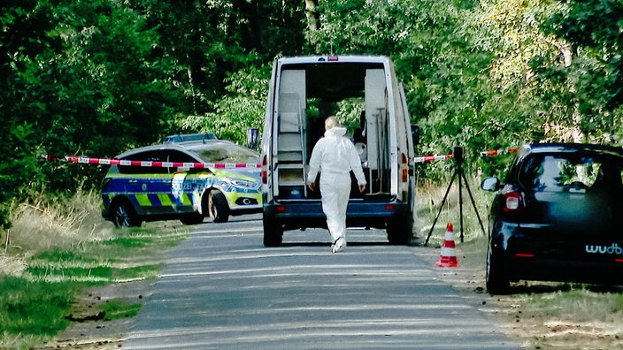 Ein abgesperrter Straßenabschnitt, an dem eine Person in weißem Schutzanzug zu einem geöffneten Transporter geht. Im Hintergrund ist ein Polizeifahrzeug und Absperrband zu sehen.