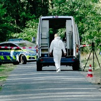 Ein abgesperrter Straßenabschnitt, an dem eine Person in weißem Schutzanzug zu einem geöffneten Transporter geht. Im Hintergrund ist ein Polizeifahrzeug und Absperrband zu sehen.