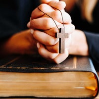 Hände einer Frau sind über einem schwarzen Buch gefaltet, über den Händen hängt ein Holzkreuz an einer Kette