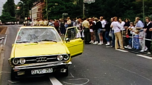 Das verunfallte Auto der Ehefrau eines Täters. Daneben stehen Polizisten und hinter einer Absperrung zahlreiche Schaulustige.