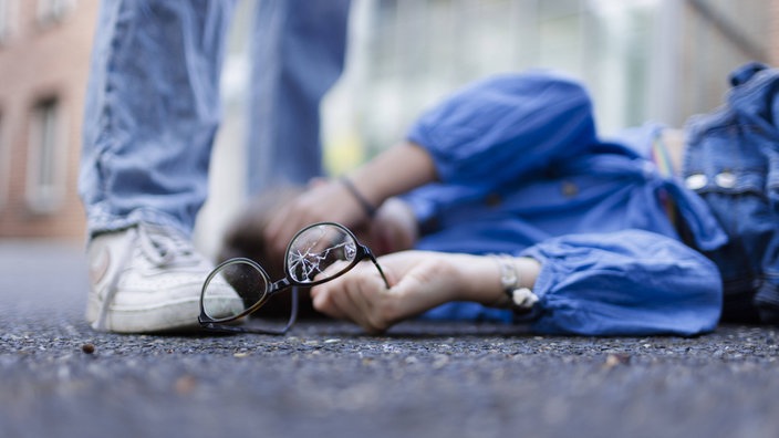Eine Person liegt auf dem Boden und hält eine kaputte Brille in der Hand. Eine andere Person steht über ihr.