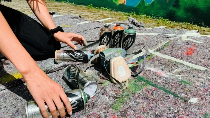 Auf dem Boden vor einer Gebäudewand liegen mehrere Spraydosen. Zwei Hände greifen danach.