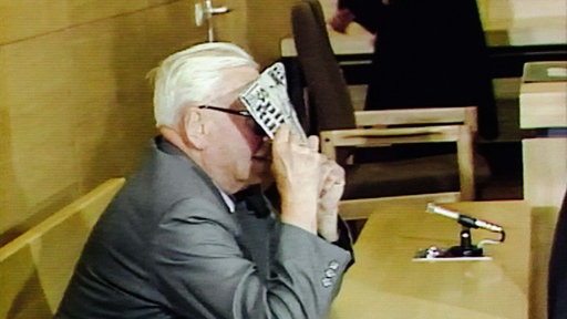 Ein älterer Mann mit Brille sitzt in einem Gerichtssaal und verdeckt sein Gesicht mit einer Bild-Zeitung.