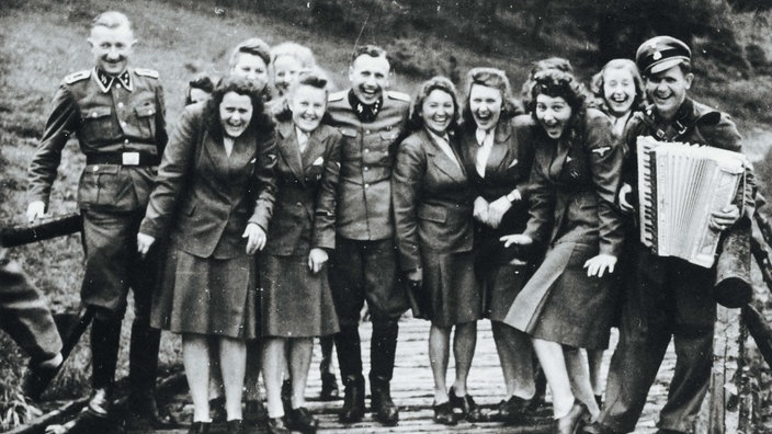 Eine Gruppe von Männern und Frauen in Uniform steht nebeneinander. Sie lachen und schauen in die Kamera. Ein Mann auf der rechten Seite spielt Akkordeon. Das Foto ist schwarz-weiß.