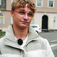Ein blonder Junge mit Brille schaut an der Kamera vorbei, im Hintergrund sind Häuser zu sehen.