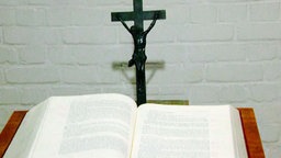 Eine aufgeschlagene Bibel liegt vor einem Kreuz Jesu.