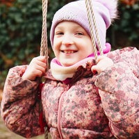 Melina, ein junges Mädchen in rosa Winterjacke und rosa Bommelmütze, sitzt auf einer Schaukel und lacht in die Kamera