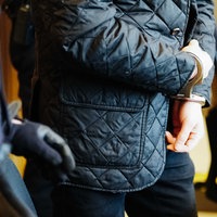 Ein Mann steht mit schwarzer Jacke und schwarzer Hose, die Hände in Handschellen, neben einem Polizisten, von beiden sieht man nur die Körpermitte