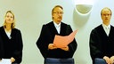 Richter Willi Erdmann steht im Gerichtssaal in einer Robe und spricht, neben ihm stehen zwei Personen in schwarzen Richterroben.