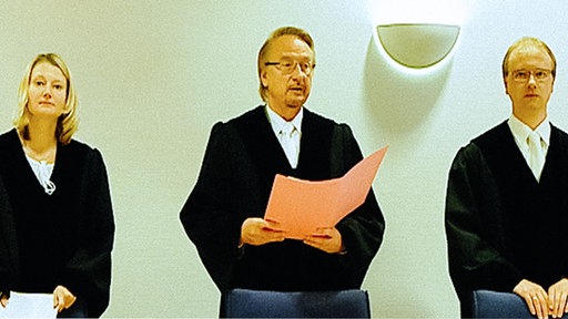 Richter Willi Erdmann steht im Gerichtssaal in einer Robe und spricht, neben ihm stehen zwei Personen in schwarzen Richterroben.