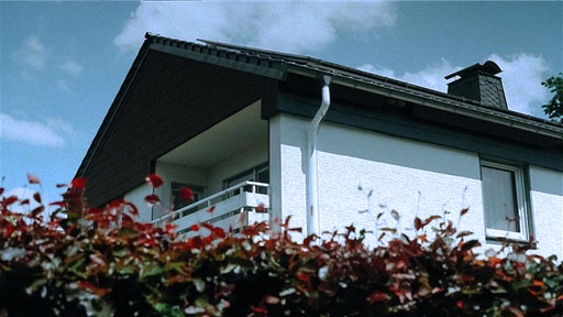 Ein weißes Haus mit schwarzem Dach und Balkon. Darunter ist eine Hecke.