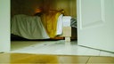 Blick durch eine geöffnete weiße Holztür auf ein Bett, auf dem eine gelbe und eine weiße Decke als kleiner Haufen liegen
