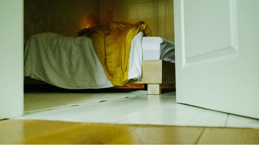 Blick durch eine geöffnete weiße Holztür auf ein Bett, auf dem eine gelbe und eine weiße Decke als kleiner Haufen liegen