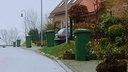 Asphaltierte Straße, am Rand der Häuser stehen grüne Mülltonnen, rechts drei Straßenlaternen, ein Auto steht in einer Einfahrt