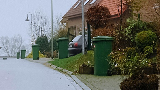 Asphaltierte Straße, am Rand der Häuser stehen grüne Mülltonnen, rechts drei Straßenlaternen, ein Auto steht in einer Einfahrt