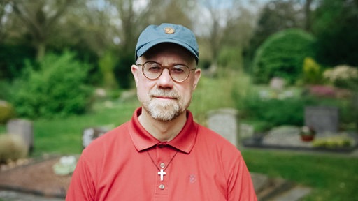 Mann mit rotem T-Shirt, Brille, blauer Cap, Kreuzkette um dem Hals